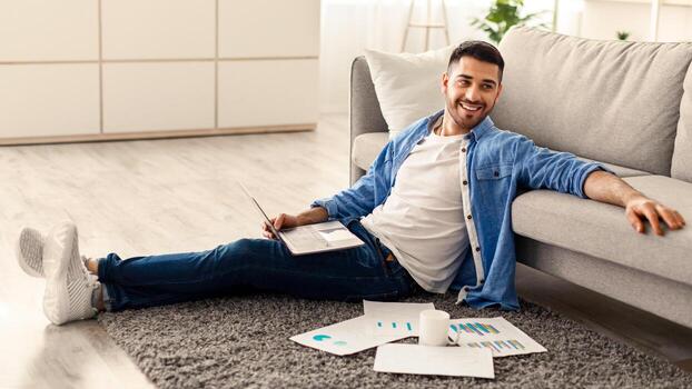 tomando descanso concepto. retrato de sonriente joven judío chico en kipá sentado en piso alfombra con computadora en su regazo, documentos y café taza, propensión en sofá y mirando lejos, descansando desde trabajo foto