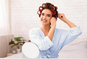 Hairstyling. Beautiful Woman Curling Hair Using Curlers Smiling To Camera Sitting At Mirror At Home. Selective Focus, Free Space photo