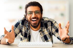 Emotional middle-eastern guy freelencer having conference with someone, sitting at worktable with computer keyboard, smiling at pc camera and cheerfully gesturing, home interior, copy space photo