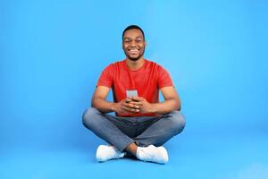 Happy african american male using cellphone smiling to camera sitting on blue studio background. Black guy using new mobile app on smartphone. People and modern gadgets concept photo