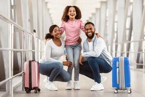 Traveling With Family. Portrait Of Cheerful Three African American People Standing In Airport Terminal With Suitcases And Hugging, Posing And Looking At Camera Waiting For Departure, Laughing photo
