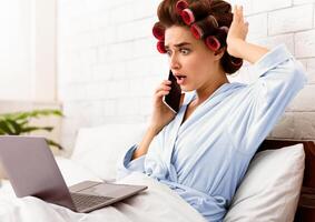 Shocked Housewife Talking On Phone Using Laptop Browsing Internet Lying In Bed At Home. Shocking News Concept photo