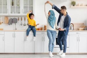Happy Islamic Family With Little Daughter Having Fun Together In Kitchen, Cheerful Middle Eastern Parents And Their Cute Female Child Dancing And Laughing, Enjoying Spending Time At Home photo