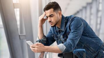 Worried young arab man looking at smartphone screen while waiting at airport terminal, frustrated middle eastern guy touching forehead, reading bad message or having problems with internet connection photo