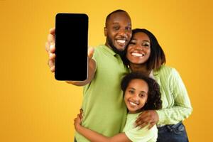 Loving african american family of three showing blank smartphone screen to camera, empty space for mockup. Excited parents and little daughter recommending mobile app, standing on yellow background photo