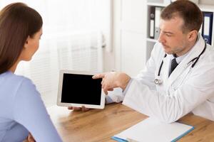 Medicine And Health Care Concept. Male doctor pointing at the screen of digital tablet with black empty screen for mockup, showing patient her test results and discussing treatment, sitting at desk photo