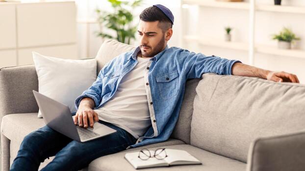 retrato de enfocado joven judío chico vistiendo kipá utilizando personal computadora sentado en sofá en vivo habitación y laboral. milenario hombre acecho , película o seminario web, comprobación correo electrónico foto