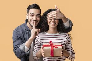 Cheerful Arab Man Covering Girlfriend's Eyes And Surprising Her With Gift, Happy Middle Eastern Guy Showing Shh Sign At Camera, Greeting Lover With Birthday Or Valentines Day, Yellow Background photo