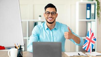 Young Arab tutor conducting online English lesson, showing thumb up, teaching foreign language from home. Guy college professior explaining grammar rules, communicating with students on web photo