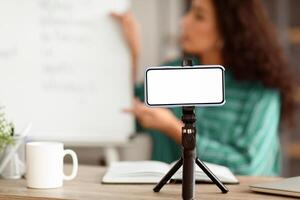 Modern Remote Education. Lady teacher shooting lecture on smartphone using tripod having conference with students, explaining material. Selective focus on blank white gadget screen, mock up photo
