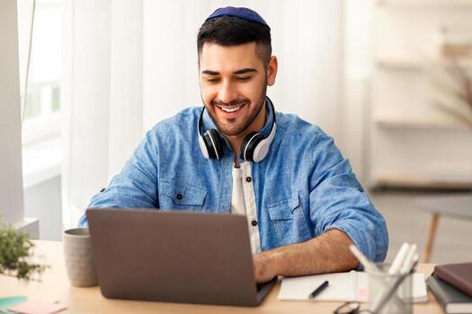 en línea educación concepto. retrato de contento judío hombre en kipa y auriculares trabajando o estudiando remotamente a hogar oficina, mirando a pantalla mecanografía en teclado, utilizando ordenador portátil y sentado a escritorio foto