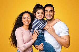 Happy Middle Easter Family Of Three With Little Daughter Embracing And Smiling At Camera, Loving Arab Parents And Cute Female Child Posing Togethet Over Yellow Studio Backgriund, Free Space photo