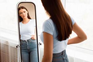Weight Gain. Unhappy Girl Touching Drooping Belly Standing In Front Of Mirror At Home. Empty Space, Selective Focus photo