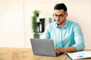 Serious young Arab guy working at his desk with laptop pc, typing on keyboard at office. Millennial Eastern businessman using portable computer, having online job, taking part in virtual meeting photo