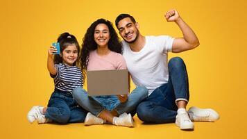 Portrait Of Happy Excited Arabic Family Of Three With Laptop And Credit Card Making Online Shopping, Middle Eastern Parents And Little Daughter Enjoying Easy Payments, Sitting Over Yellow Background photo