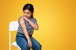Covid-19 Vaccination. Vaccinated Little Arab Girl Looking At Arm With Plaster After Vaccine Injection, Cute Middle Eastern Female Kid Sitting On Chair Over Yellow Studio Background, Copy Space photo