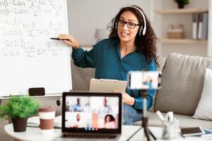 Videoconference And Training. Class of diverse students learning remotely online using web app. Lady teacher in headset providing lesson looking at pc screen, recording on cellphone on tripod photo