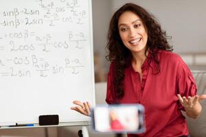 Smiling lady sitting in front of smartphone on stabilizer, recording or having live videoconference with students. Happy young smart woman filming educational lecture, explaining theme photo