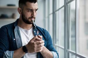 Bad habits, tobacco addiction, stress and suffering from depression. Frustration millennial attractive bearded guy lights cigarette with lighter looking out large window background, copy space photo