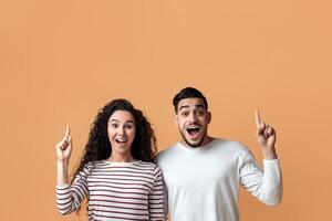 Idea Concept. Joyful Middle Eastern Couple Pointing Fingers Up And Looking At Camera, Young Excited Arab Spouses Found Problem Solution, Having Inspiration Moment While Posing On Yellow Background photo