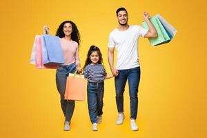Happy Cheerful Arab Parents And Their Little Daughter With Bright Shopping Bags Walking Over Yellow Studio Background, Joyful Middle Eastern Family Of Three Carrying Purchases And Smiling, Copy Space photo