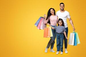 Seasonal Sales. Portrait Of Happy Arab Family With Little Daughter Holding Lots Of Bright Shopping Bags, Middle Eastern Parents And Child Posing Over Yellow Background In Studio, Copy Space photo