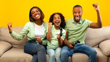 Family time activities. Happy black parents and their daughter playing games while sitting on sofa over yellow background. Dad, mother and child holding joysticks, winning game photo