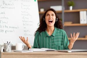 Fatigued From Work. Tired young teacher in headset screaming out loud, gesturing and looking up, having lesson with students, tutoring English language online, sitting at desk and yelling photo