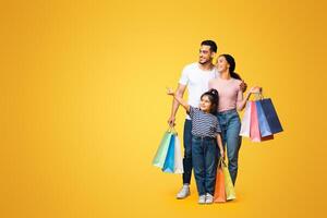 Family Shopping. Cute Little Girl Pointing Finger Aside Showing Something To Her Arab Parents While They Standing Together With Shopper Bags Over Yellow Studio Background, Panorama With Copy Space photo