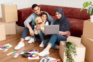 Relocation Concept. Joyful Middle Eastern Family Of Three People Using Laptop Sitting On Floor Among Moving Boxes And Paint In New House, Choosing And Ordering Furniture, Discussing Decor And Design photo