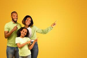 Special offer. Positive african american family of three pointing fingers aside at free space and smiling at camera on yellow background, presenting empty place for advert, banner photo