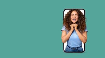 A woman with curly hair joyfully interacts with a large smartphone screen, showcasing excitement and emotion, symbolizing modern communication and connection through technology. photo