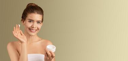 A woman enjoys her skin care routine, applying cream while smiling. She is isolated in a cozy space with a simple background, highlighting her pampering moment. photo
