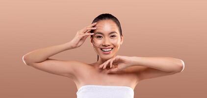 Asian woman enjoys her skincare routine, applying products to her face with a smile. She showcases the importance of self-care in a relaxed, isolated setting with ample empty space. photo