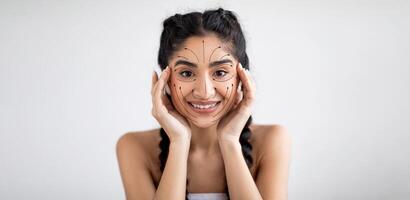 Indian woman is happily engaging in her skincare routine, applying products with care. She has a bright smile and is surrounded by an empty, well-lit space for her self-pampering session. photo