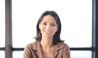 A woman with a cheerful expression sits in a well-lit environment as facial recognition technology analyzes her features. The background is plain, emphasizing her face. photo