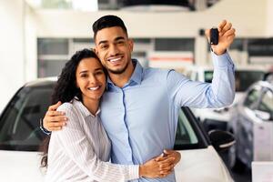 Happy young family buying new car in luxury showroom, standing buy brand new white auto, embracing and showing automatic key, copy space. Cheerful couple bought nice auto, car store interior photo