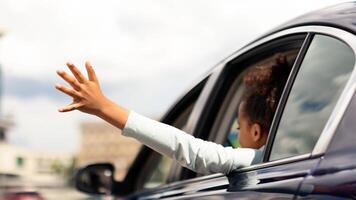 African American Kid Girl Putting Arm Out Of The Car Window Having Fun While Riding With Family In City. Summer Road Trip Adventure Concept. Back View, Selective Focus photo