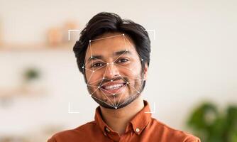 A young man with a cheerful expression stands in a well-lit indoor environment. Facial recognition technology highlights his face, indicating advancements in identification and security. photo