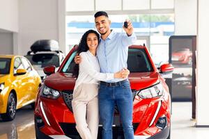Happy beautiful loving muslim family young arab man and long-haired woman buying new car in salon, standing over red luxury automobile, hugging and showing key from their new auto, copy space photo