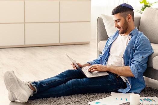 persona de libre dedicación jornada laboral. sonriente milenario hombre empleado estudiante trabajando estudiando desde hogar sentado en piso alfombra en vivo habitación. masculino utilizando ordenador portátil comprometido en electrónico papeleo haciendo distante tarea foto