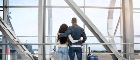 Traveling Together. Rear View Of Young Couple Standing At Airport Terminal, Romantic Lovers Embracing And Looking Out Of Window While Waiting For Flight, Ready To Honeymoon Trip, Free Space photo