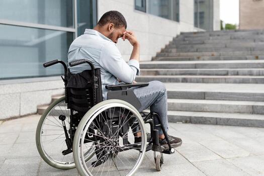 Disability and accessible environment problems. Upset handicapped black man in wheelchair in front of stairs without ramp, having no possibility to enter building, copy space photo