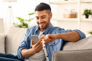 Useful App. Handsome Israeli Man In Kippah Using Smartphone While Relaxing On Couch At Home, Jewish Guy Sitting On Sofa In Living Room, Browsing Internet Or Messaging With Friends, Free Copy Space photo