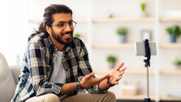 Handsome arab guy blogger streaming from home, sitting on sofa, looking at mobile phone camera, talking, gesturing, smiling. Indian guy blogger having stream, chatting with followers, copy space photo
