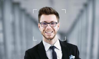A young man with glasses and a confident smile stands in a modern setting. Facial recognition technology is applied to his face, highlighting its features against a blurred background. photo