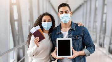 Middle Eastern Couple In Protective Medical Masks Showing Blank Digital Tablet At Camera While Standing Together At Airport Terminal With Passports And Tickets In Hands, Travelling During Pandemic photo