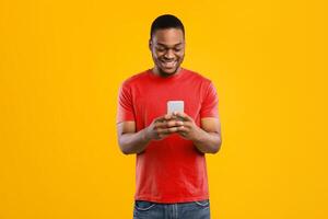 Smiling Black Man Using Smartphone Browsing Internet And Scrolling Social Media News Feed Standing Over Yellow Studio Background. Millennial Guy Using New Mobile Application. People And Gadgets photo