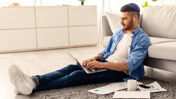 Serious millennial jewish man using laptop sitting on floor carpet leaning on couch, focused casual guy communicating online, writing emails, distantly working or studying on computer doing paperwork photo