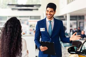 Wealthy middle-eastern young woman choosing new car, having conversation with friendly sales manager at brand new vip showroom, dealer pointing at yellow comfy auto and smiling, side view photo